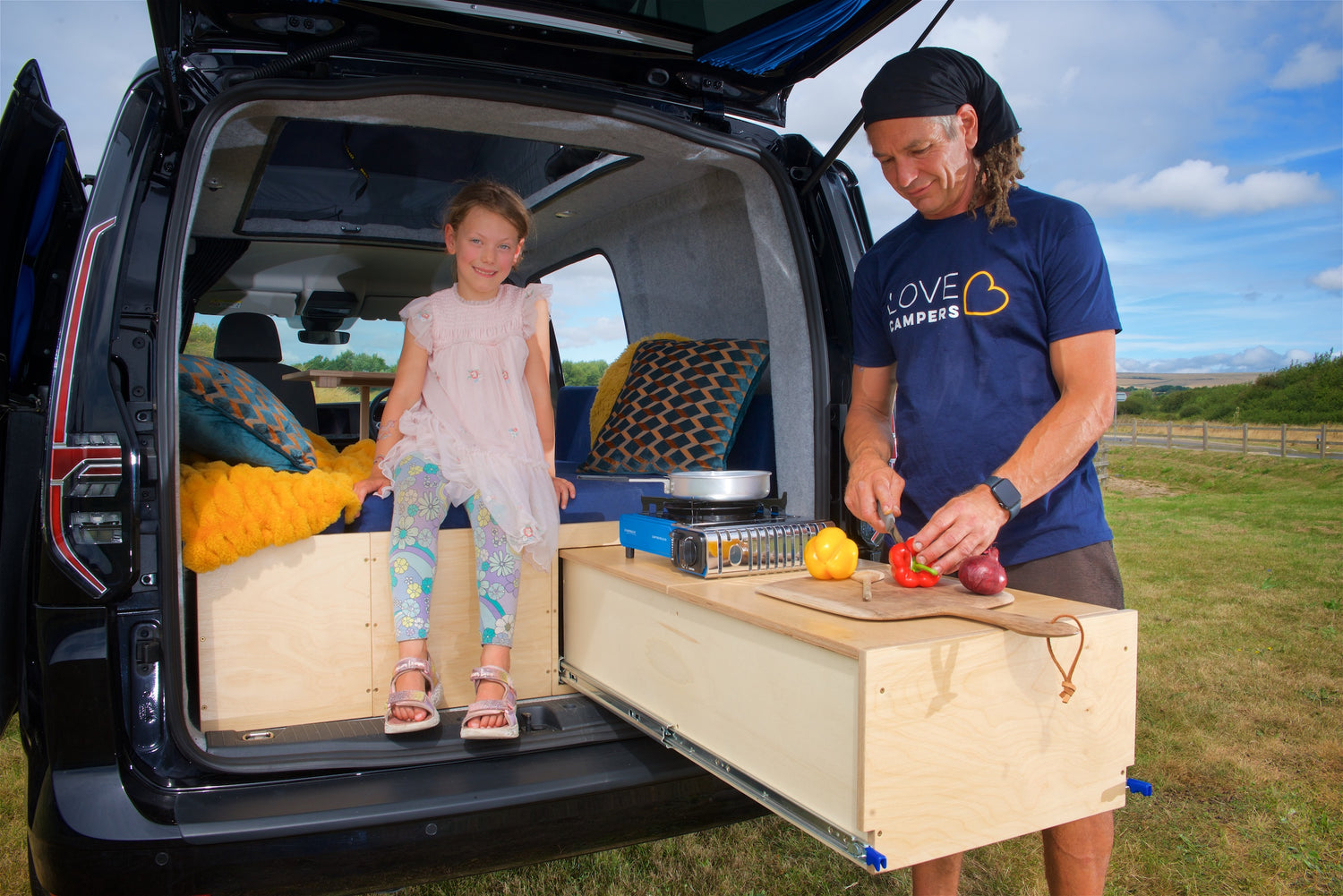 A man chops vegetables in the Sunbox kitchen area with a child sat inside the van